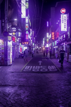 A vibrant urban street scene at night, illuminated by numerous neon signs with Korean characters. The street is bustling with people walking amidst a backdrop of colorful lights and signs. The ground has visible textures and is marked by a painted arrow and Korean text.