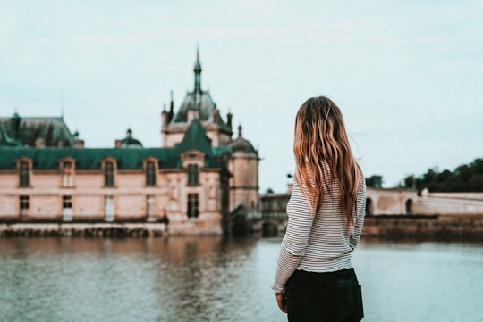 woman facing beige and green concrete building and body of water in Château de Chantilly France