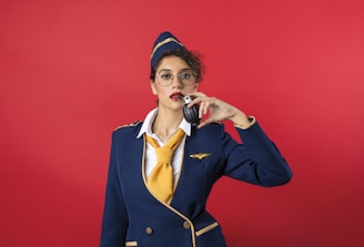 A person wearing a flight uniform with a navy blue jacket and a yellow tie holds a black object resembling a grenade. The individual has curly hair, wears glasses, and poses against a solid red background.