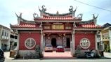 A traditional Chinese temple with ornate architecture featuring intricate roof designs and vibrant decorations. The façade is adorned with red bricks and golden embellishments. At the entrance, there are statues of lions on either side, and vehicles are parked in front, including a car and bicycles. The temple is surrounded by buildings that have a colonial architectural style.