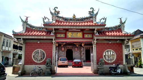 A traditional Chinese temple with ornate architecture featuring intricate roof designs and vibrant decorations. The façade is adorned with red bricks and golden embellishments. At the entrance, there are statues of lions on either side, and vehicles are parked in front, including a car and bicycles. The temple is surrounded by buildings that have a colonial architectural style.