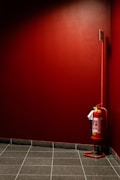 A red fire extinguisher is mounted against a red-walled corner. The floor is covered with black and gray tiles, and there's a small sign attached to the extinguisher.