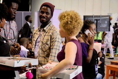 A group of people are gathered in a busy indoor setting, possibly at a conference or event. Several individuals are holding Krispy Kreme doughnut boxes, engaged in conversation. One person is talking on a mobile phone while others are interacting with each other. The atmosphere appears lively and bustling.