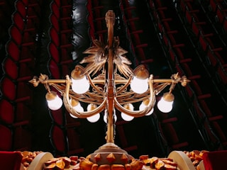 An ornate chandelier with multiple glowing bulbs is hanging in a luxurious interior. The structure features intricate golden designs and motifs, with a dark background revealing rows of red cushioned theater seats.