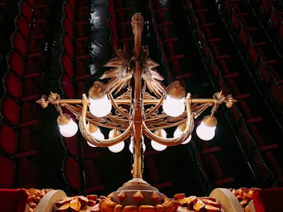 An ornate chandelier with multiple glowing bulbs is hanging in a luxurious interior. The structure features intricate golden designs and motifs, with a dark background revealing rows of red cushioned theater seats.