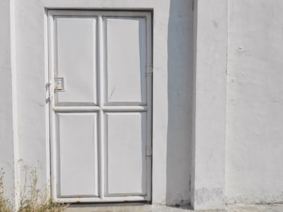 A white metal door with a slightly worn texture and four rectangular panels is set in a concrete wall. The door has a simple square handle and is surrounded by subtle cracks in the wall. Some light vegetation is visible at the base, suggesting an outdoor setting.