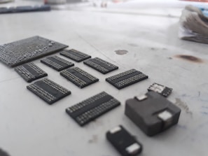 A collection of various microchips and electronic components neatly arranged on a light-colored surface. The components appear to be of different shapes and sizes, with visible metallic connection points. The focus is on the chips, giving them a detailed and textured look, while the background is blurred.