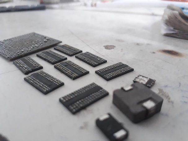 A collection of various microchips and electronic components neatly arranged on a light-colored surface. The components appear to be of different shapes and sizes, with visible metallic connection points. The focus is on the chips, giving them a detailed and textured look, while the background is blurred.