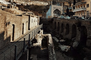 A historical site featuring old brick buildings, arches and a walkway, with a mix of domed roofs. There are cars parked near a Gothic-style building with decorative windows and a detailed facade. The scene is set against a backdrop of hills and vegetation.