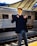A young person wearing reflective sunglasses, a dark blazer, and jeans stands confidently on a train platform. The background features a sleek metallic train carriage and a modern station roof structure.