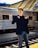 A young person wearing reflective sunglasses, a dark blazer, and jeans stands confidently on a train platform. The background features a sleek metallic train carriage and a modern station roof structure.