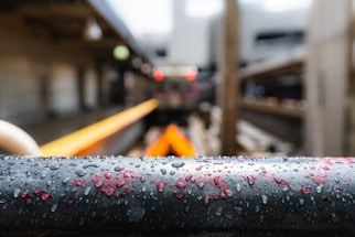 A close-up of a wet black metal railing with droplets of water and patches of red paint. The background is blurred, showing what appears to be an industrial setting with metal beams and pipes, and a hint of an orange-colored object.