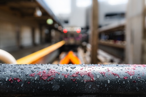 A close-up of a wet black metal railing with droplets of water and patches of red paint. The background is blurred, showing what appears to be an industrial setting with metal beams and pipes, and a hint of an orange-colored object.