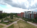 A modern urban landscape featuring a mix of residential buildings and green spaces. The scene includes contemporary apartment buildings with red brick and white facades. Well-maintained gardens, trees, and walking paths create a peaceful environment. The sky is cloudy with patches of blue.