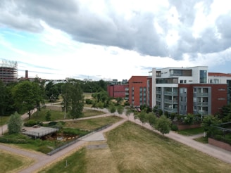 A modern urban landscape featuring a mix of residential buildings and green spaces. The scene includes contemporary apartment buildings with red brick and white facades. Well-maintained gardens, trees, and walking paths create a peaceful environment. The sky is cloudy with patches of blue.