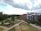 A modern urban landscape featuring a mix of residential buildings and green spaces. The scene includes contemporary apartment buildings with red brick and white facades. Well-maintained gardens, trees, and walking paths create a peaceful environment. The sky is cloudy with patches of blue.