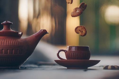 A terracotta teapot and cup set on a table, with heart-shaped cookies seemingly dropping into the cup. The background is softly blurred with warm, natural tones, suggesting an outdoor setting or an area with incoming sunlight through windows.