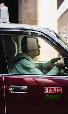A man sits in the driver’s seat of a classic taxi, wearing a formal white shirt. Sunlight streams through the window, casting reflections on the vehicle's maroon exterior. The taxi sign on the car door includes Japanese script along with the word 'taxi'.