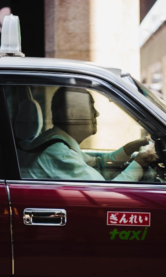 A man sits in the driver&rsquo;s seat of a classic taxi, wearing a formal white shirt. Sunlight streams through the window, casting reflections on the vehicle's maroon exterior. The taxi sign on the car door includes Japanese script along with the word 'taxi'.