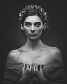 A monochrome portrait of a woman with a serious expression. She wears a floral headband and holds her robe off her shoulders, highlighting her collarbones. The lighting casts soft shadows, adding a dramatic effect to the image.