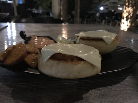 A close-up of a plate containing two round bread rolls topped with melted cheese, accompanied by thick-cut potato wedges and a side of creamy sauce with a small portion of coleslaw. The setting is dimly lit, contributing to a cozy dining atmosphere.