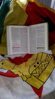 An open book, possibly a religious text, placed on a colorful fabric that has yellow, red, green, and black sections. The fabric features a graphic design with geometric patterns.