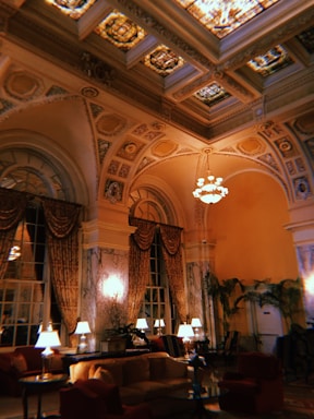 An opulent, elegantly decorated room featuring high, ornate ceilings with intricate designs and stained glass panels. The room is adorned with luxurious drapes and multiple seating areas with plush furniture. Warm lighting from lamps creates a cozy atmosphere, complemented by decorative plants scattered throughout the space.