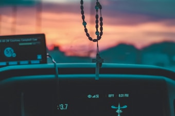 A hanging rosary with a cross is prominently displayed inside a vehicle, silhouetted against a vibrant sunset. Dashboard displays show various vehicle metrics, including temperature and time. The background features a blurred landscape, creating a peaceful and contemplative atmosphere.
