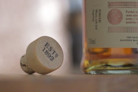 A close-up view of a cork with the inscription 'ESTD 1892' lying on its side next to a bottle labeled 'The Balvenie'. The label includes details about the whiskey, and the scene is softly lit, giving it a warm, inviting feel.