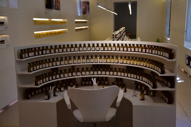 A stylish interior with a curved display shelf filled with rows of small uniform dark glass bottles, each topped with a stopper. The bottles are arranged meticulously, occupying several tiers of the shelving unit. A modern white chair is placed in front of the shelves, and a few small implements or droppers are on the lower tier. Soft lighting highlights the orderly arrangement, set against light-colored walls and a glossy tiled floor.