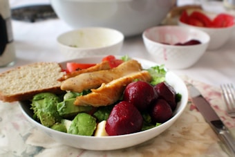 A bowl filled with vibrant green avocado slices, roasted chicken pieces, red beets, and leafy greens, accompanied by a slice of bread. The setting includes more bowls in the background, suggesting a meal setup.