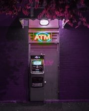 An ATM machine is located on a dark purple brick wall under a neon sign. The neon sign above the machine is colorful and reads 'ATM' in bright red, green, and blue lights. The area is dimly lit with purplish hues, creating a moody atmosphere.