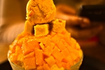 A close-up of a dessert featuring a mound of shaved ice or similar base, topped with fresh mango cubes. There is a scoop of mango ice cream on top, adding to the tropical theme. The background is blurred, focusing attention on the dessert.