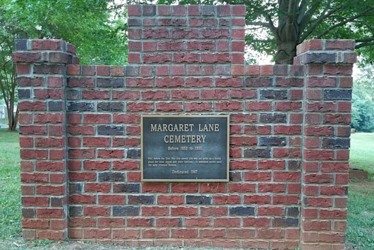 A brick wall with a plaque bearing the text 'Margaret Lane Cemetery'. The wall is surrounded by grass and trees, with visible greenery in the background.