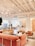 A modern and cozy interior space with a warm color palette. The room features plush pink and beige seating arrangements, including sofas and armchairs. There is a large, stylish chandelier hanging from the white ceiling with exposed beams. The space is open and airy with large windows allowing natural light to flood in. A reception desk is present in the background with a person sitting at it. There are also small tables with decorative plants and books scattered throughout the area, contributing to a welcoming atmosphere.