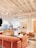 A modern and cozy interior space with a warm color palette. The room features plush pink and beige seating arrangements, including sofas and armchairs. There is a large, stylish chandelier hanging from the white ceiling with exposed beams. The space is open and airy with large windows allowing natural light to flood in. A reception desk is present in the background with a person sitting at it. There are also small tables with decorative plants and books scattered throughout the area, contributing to a welcoming atmosphere.