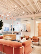 A modern and cozy interior space with a warm color palette. The room features plush pink and beige seating arrangements, including sofas and armchairs. There is a large, stylish chandelier hanging from the white ceiling with exposed beams. The space is open and airy with large windows allowing natural light to flood in. A reception desk is present in the background with a person sitting at it. There are also small tables with decorative plants and books scattered throughout the area, contributing to a welcoming atmosphere.