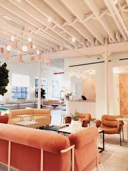 A modern and cozy interior space with a warm color palette. The room features plush pink and beige seating arrangements, including sofas and armchairs. There is a large, stylish chandelier hanging from the white ceiling with exposed beams. The space is open and airy with large windows allowing natural light to flood in. A reception desk is present in the background with a person sitting at it. There are also small tables with decorative plants and books scattered throughout the area, contributing to a welcoming atmosphere.