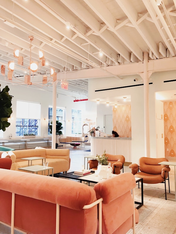 A modern and cozy interior space with a warm color palette. The room features plush pink and beige seating arrangements, including sofas and armchairs. There is a large, stylish chandelier hanging from the white ceiling with exposed beams. The space is open and airy with large windows allowing natural light to flood in. A reception desk is present in the background with a person sitting at it. There are also small tables with decorative plants and books scattered throughout the area, contributing to a welcoming atmosphere.