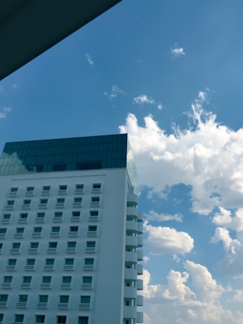A modern high-rise building with numerous windows and a rooftop section with glass panels stands against a backdrop of a bright blue sky filled with scattered white clouds. The building appears to have a sleek design with curved balconies visible.