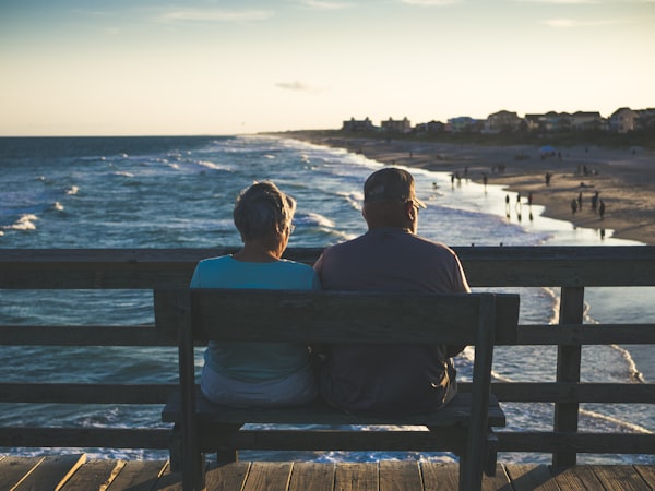 Deux personnes âgées portant des T-shirts bleu et violet assises sur un banc en bois face à la mer et à la plage