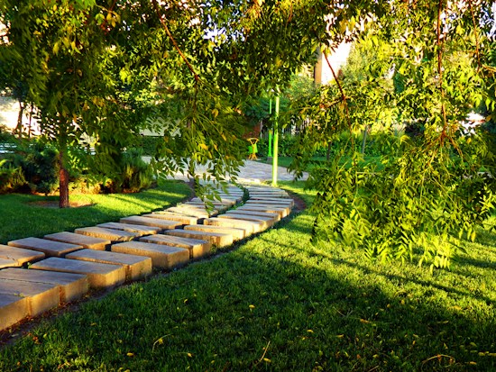 A serene garden scene with a stone pathway leading through lush, green grass. Overhanging tree branches dapple the area with sunlight, casting intricate shadows. The bright sunlight highlights the vibrant greenery, creating a peaceful and inviting atmosphere.