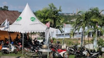 A scene featuring a tent with a logo and the words 'PT. Kaltim Parna Industri' printed on it. Several motorcycles are parked under and around the tent. The background shows a landscape with palm trees, a building, and industrial structures in the distance.
