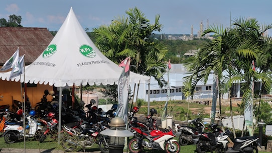 A scene featuring a tent with a logo and the words 'PT. Kaltim Parna Industri' printed on it. Several motorcycles are parked under and around the tent. The background shows a landscape with palm trees, a building, and industrial structures in the distance.