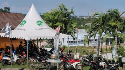 A scene featuring a tent with a logo and the words 'PT. Kaltim Parna Industri' printed on it. Several motorcycles are parked under and around the tent. The background shows a landscape with palm trees, a building, and industrial structures in the distance.