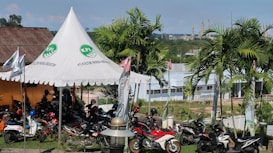 A scene featuring a tent with a logo and the words 'PT. Kaltim Parna Industri' printed on it. Several motorcycles are parked under and around the tent. The background shows a landscape with palm trees, a building, and industrial structures in the distance.