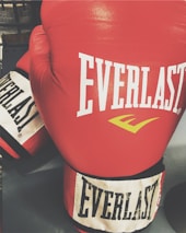 A pair of red boxing gloves with the brand name 'Everlast' prominently displayed on them. The gloves have white and black colored cuffs, and the surface appears glossy, suggesting they are made of leather.