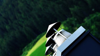 A chimney with a metallic cap stands in the foreground against a blurred backdrop of lush green foliage and a field. The angle gives a dynamic perspective, emphasizing the architectural structure.