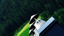 A chimney with a metallic cap stands in the foreground against a blurred backdrop of lush green foliage and a field. The angle gives a dynamic perspective, emphasizing the architectural structure.