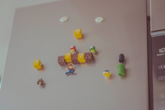 A collection of small, colorful Super Mario-themed magnets is attached to a refrigerator surface. These include Mario, question blocks, mushrooms, and a green pipe, resembling elements from the iconic video game series.